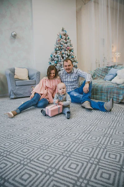happy family new year photo session: mother, father and little child are sitting near the new year tree in a bright interior 1