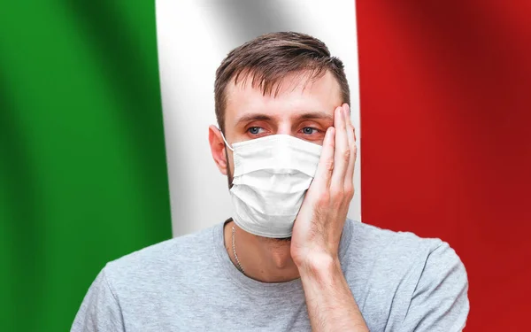 man on the background of the Italian flag in a medical mask on his face that protects against coronavirus and other diseases, holds in his hands a white blank piece of paper for your text