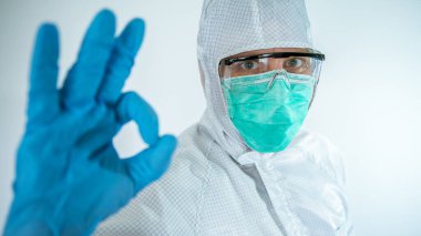 Kendine güvenen beyaz bir erkek doktorun yakın plan portresi koruyucu bir takım elbise ve cerrahi maske takıyor ve beyaz arka planda izole edilmiş baş parmakları gösteriyor. Biyolojik tehlike koronavirüsü Covid-19