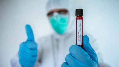 Koruyucu giysisi olan bir erkek bilim adamı. COVID-19 için negatif kan tübü olan ve baş parmağını veren beyaz bir adam. Sonuç iyi. Coronavirus için aşı araştırması