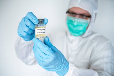Kafkas bir doktor laboratuarda, koronavirüs hastalıkları için sıvı aşı tutuyor. Covid-19 aşısıyla koruyucu kıyafet giyen bir erkek bilim adamı. Tıbbi ve sağlık konsepti, biyolojik tehlike.