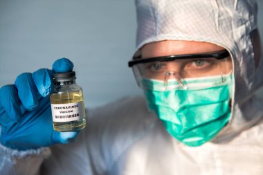 Kafkas bir doktor laboratuarda, koronavirüs hastalıkları için sıvı aşı tutuyor. Covid-19 aşısıyla koruyucu kıyafet giyen bir erkek bilim adamı. Tıbbi ve sağlık konsepti, biyolojik tehlike.