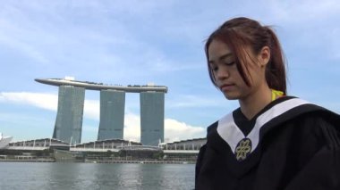 4k, Asyalı kadın Singapur limanında mezuniyet şapkasını taktı ve arka planda Marina Bay Sands lüks oteli olan kameraya baktı.
