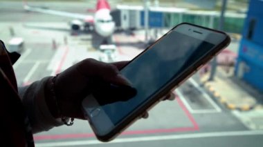 Uluslararası havaalanında uçak geçmişi olan 4K Genç kadın cep telefonu tutuyor. Asyalı kız akıllı telefonu uçakla kontrol ediyor. Gezginler uçak yolculuğu için terminalde bekliyorlar.