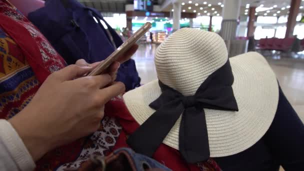 4K年轻女子在国际机场用手机打字 亚洲女孩拿着帽子检查智能手机 人们坐在候机室门口等着坐飞机去旅行 — 图库视频影像