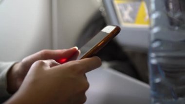 4K, Asyalı kadın uçuş sırasında elinde akıllı telefon tutuyor. Genç gezgin uçaktaki bir kısa mesajı görmek için telefon cihazını kullanarak uçakta oturuyor. Turistler taze su şişesiyle seyahat ediyorlar.