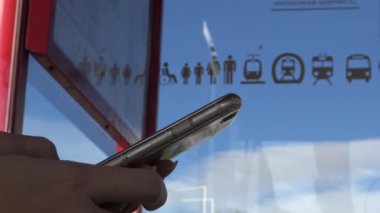 4K Genç Asyalı kadın akıllı telefonuyla SMS yazıyor, Madrid 'de bir güneş gününde otobüs durağında beklerken telefonları kontrol ediyor. İnternette sörf yapmak için mobil bir cihaz kullanmak ve sosyal ağları kontrol etmek - Dan