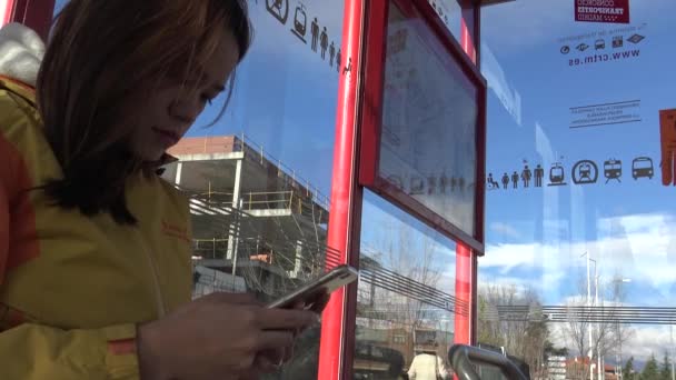 아시아인 스마트폰으로 Sms 문자를 마드리드 거리의 햇살을 받으며 정류장에서 기다리는 — 비디오
