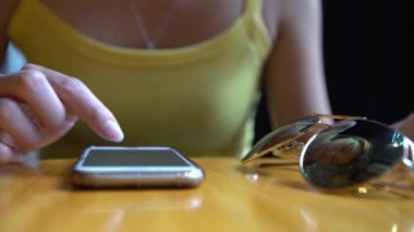 Kahvehane masasında akıllı telefon kullanan 4K Asyalı kadın. Sohbet etmek ve insanlarla iletişim kurmak için ekrana dokun. İnternette sörf yapmak için iyi bir zaman, güneş gözlüğü yakınındaki telefon cihazından sosyal ağları kontrol ediyor.