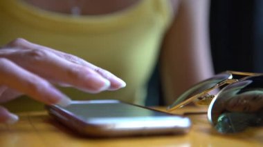 Kahvehane masasında akıllı telefon kullanan 4K Asyalı kadın. Sohbet etmek ve insanlarla iletişim kurmak için ekrana dokun. İnternette sörf yapmak için iyi bir zaman, güneş gözlüğü yakınındaki telefon cihazından sosyal ağları kontrol ediyor.