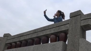 4K Asyalı kadın geleneksel mimari köprüsü olan Çin Bahçesi 'nde cep telefonu kullanıyor. İnternette sörf yapmak, telefon üzerinden yapılan sosyal ağları kontrol etmek, resimleri görmek için akıllı telefon kullanmak - Dan