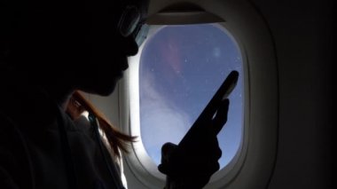 4K, Asyalı kadın uçuş sırasında pencerenin yanında akıllı telefon tutuyor. Genç gezgin, uçakta bir mesaj görmek için telefon cihazını kullanarak uçağın içine oturdu. Seyahat eden turistler - Dan