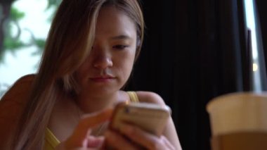 Kahvehanede akıllı telefon kullanan 4K Asyalı kadın. Rahatlamak ve kahvenin tadını çıkarmak için iyi bir zaman. İnternette sörf yapıyor, sosyal ağ sohbetlerini kontrol ediyor ve telefon cihazı Dan ile insanlarla bağlantı kuruyor.