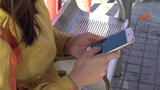 4K Jeune femme asiatique écrit du texte SMS avec smartphone, vérifie le téléphone pendant l'attente dans l'arrêt de bus à une journée de soleil dans la rue Madrid. Utilisation d'un appareil mobile pour surfer sur Internet et contrôle les réseaux sociaux Dan 