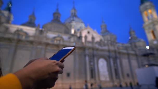 アジアの女性は夜の背景に柱の聖母大聖堂とスマートフォンを使用しています サラゴサ ダンの有名なランドマークで旅行中にデバイスのモバイルを使用している人々 — ストック動画