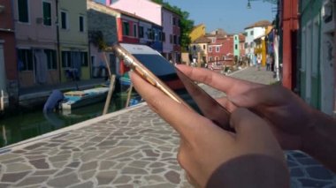 4K, Asyalı kadın Burano adasının güzel ve renkli evleri olan caddesinde geziyor ve akıllı telefon kullanıyor. Yazın bulanık kanalı ve teknesi olan bir cep telefonu tutan kişi - Dan
