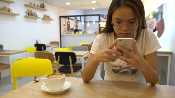 4K亚洲女孩与眼镜 看着她的智能手机聊天和联系人们在咖啡店 在网上冲浪 查看社交网络 在手机上发短信的好时机 — 图库视频影像