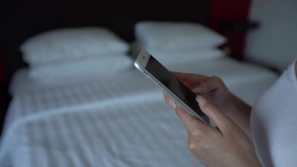 4Kアジアの女性は ベッドの上に白いシーツでホテルの部屋の電話機を手に持っています インターネットサーフィンのためのスマートフォンを使用して ソーシャルネットワークをチェックし 写真を参照してください — ストック動画