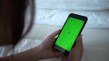 4K, yatakta yatan güzel Asyalı kadın beyaz çarşaflı bir otel odasında yeşil ekran telefon gösteriyor. Akıllı telefonu bir reklam için kullanmak. Kız tutma aygıtı - Dan