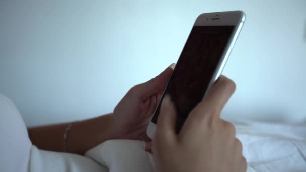 4Kアジアの女性が白いシーツでベッドの上に横になり 美しいホテルの部屋でスマートフォンを使用しています インターネットをサーフィンし ソーシャルネットワークをチェックし 彼女の携帯電話デバイス上の写真を参照してください — ストック動画