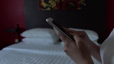 Yavaş hareket eden Asyalı kadın, otel odasındaki beyaz çarşaflı telefon cihazını elinde tutuyor. İnternette sörf yapmak için akıllı telefonu kullanmak, sosyal ağları kontrol etmek ve resimlere bakmak.