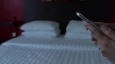 Yavaş hareket eden Asyalı kadın, otel odasındaki beyaz çarşaflı telefon cihazını elinde tutuyor. İnternette sörf yapmak için akıllı telefonu kullanmak, sosyal ağları kontrol etmek ve resimlere bakmak.