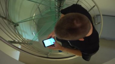 4K, beyaz adam akıllı telefon kullanıyor ve evde merdivenlerden çıkıyor. İç evdeki modern cam merdivenlerde ekran telefonuna dokunuyorum. İnternette sörf yapmak, sosyal ağı kontrol etmek ve metin-Dan yazmak
