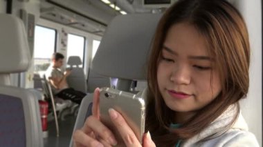 4K Gülümseyen Mutlu Asyalı Kadın İnsanlar Arkaplanda Otururken Cep Telefonu Kullanarak Trende Bekliyor. İnternette sörf yapmak, sosyal ağı kontrol etmek ve akıllı telefondan Dan 'e metin yazmak