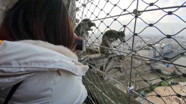パリの背景を持つノートルダム大聖堂の美しいガーゴイル像の写真を撮るためにスマートフォンを使用して観光客の女性 休暇でカメラの携帯電話で写真を撮る人々 — ストック動画