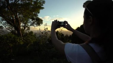 4K Asyalı kadın dağda yürüyüş yapıyor ve Taipei şehrinin ufuk çizgisine telefonla fotoğraf çekiyor. Taipei 101 binası Dahu Park 'taki dağdan yükselen diğer binaların çok üstünde.