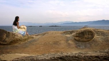 4K Güzel Asyalı kadın plajdaki kayada oturuyor, New Taipei City, Tayvan-Dan 'deki Yehliu Geopark' ın kayalık kıyı manzarasına bakıyor.