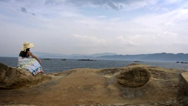 4K美しいアジアの若い女性が海岸の岩の上に座って 新北市のYehuliuジオパークの野生の岩の海岸風景を見てダン — ストック動画
