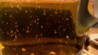 4K, taze soğuk birayı kapat. Cam yüzeyine su damlaları, saydam sarı arkaplan. Bir sürü altın bira köpüğü. Bardaki ahşap bir masada bir kupa bira.