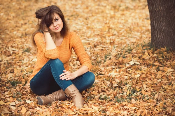 Fashion portrait of a girl in jeans and a sweater in the nature sitting on yellow autumn leaves — Stock Photo, Image