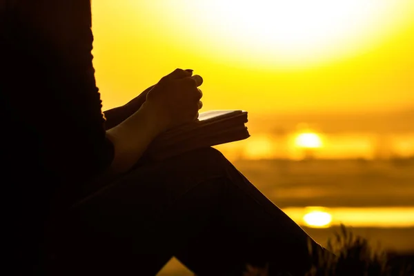 Silhueta das mãos da mulher orando a Deus na natureza witth a Bíblia ao pôr do sol, o conceito de religião e espiritualidade — Fotografia de Stock