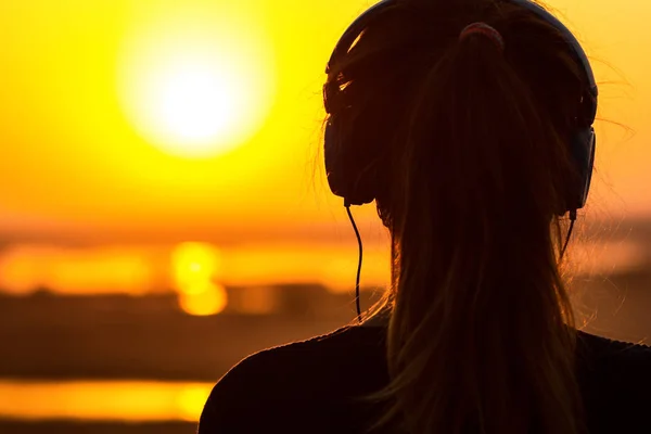 Silhouet van een vrouw in de koptelefoon, het meisje die genieten van muziek op de zonsondergang — Stockfoto