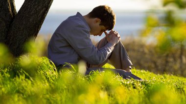 young man praying to God in the nature bowing his head to his knees, concept religion clipart