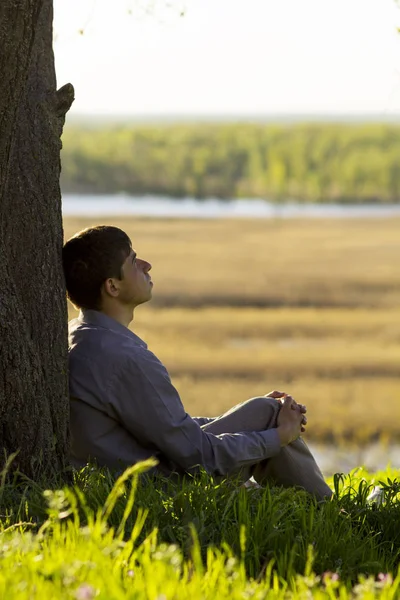 Joven sentado cerca del árbol y meditando, el hombre pasa su tiempo en la naturaleza — Foto de Stock