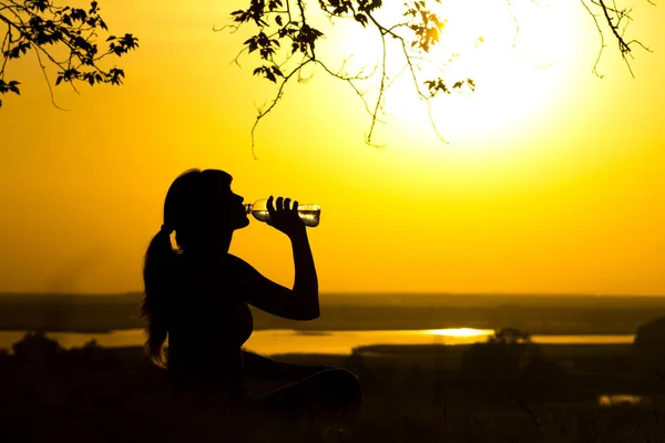 Silhouette di una donna che beve acqua dopo un allenamento di fitness in natura, profilo femminile al tramonto, concetto di sport e relax — Foto Stock