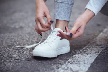 Kız asfalt yolda beyaz spor ayakkabılara ayakkabı bağlamak için çömelmiş, sonbahar sporu kavramı açık havada.