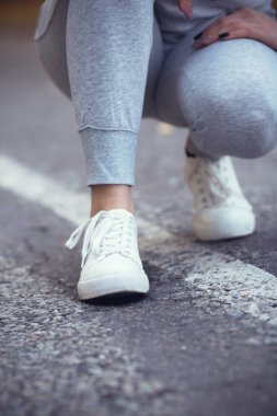 Kız asfalt yolda beyaz spor ayakkabılara ayakkabı bağlamak için çömelmiş, sonbahar sporu kavramı açık havada.