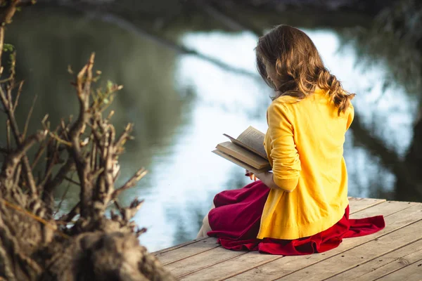 Bella giovane donna sulle rive del fiume seduta su una piattaforma di legno con un libro, adolescente che legge letteratura di finzione sulla natura, hobby concetto — Foto Stock