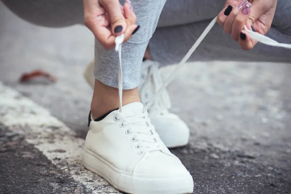 3.女孩子蹲在地上把鞋带系在柏油路的白运动鞋上,这是秋日户外运动的概念 — 图库照片