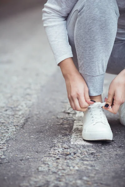 Chica sentada en cuclillas para atar cordones de zapatos en zapatillas blancas en asfalto carretera, otoño deporte concepto al aire libre — Foto de Stock