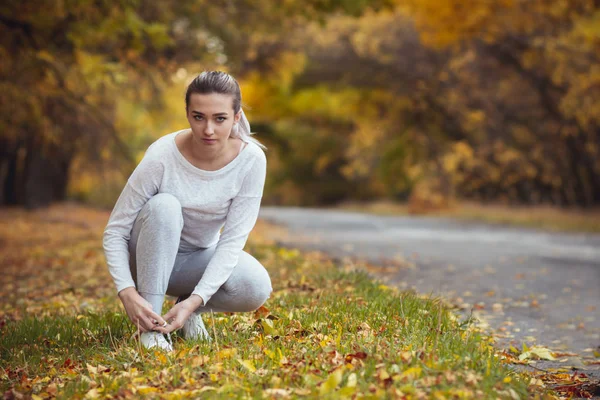 Mädchen hockt auf weißen Turnschuhen auf Asphaltstraße und bindet Schnürsenkel, Herbstsportkonzept im Freien — Stockfoto
