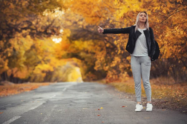 Junge Frau steht und wartet bei kaltem Herbstwetter auf einer asphaltierten Straße, ein Mädchen versucht, mit einer Handbewegung von einer Vorstadtautobahn zu kommen — Stockfoto