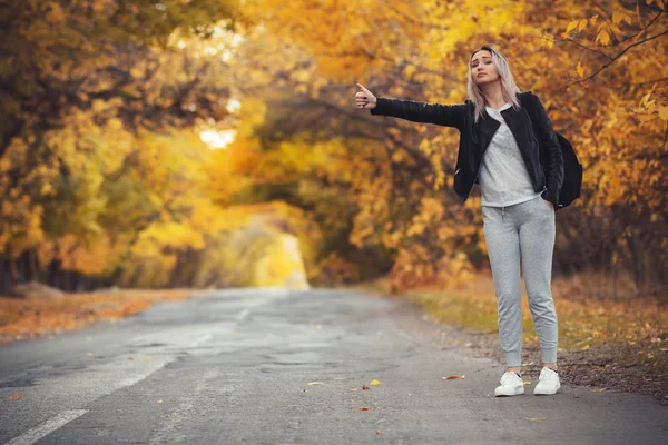 若い女性は寒い秋の天候のアスファルトの道路に立って待っている女の子は手のジェスチャーを示すヒッチハイカーによって郊外の高速道路から取得しようとします — ストック写真