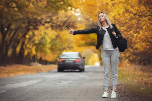 Junge Frau steht und wartet bei kaltem Herbstwetter auf einer asphaltierten Straße, ein Mädchen versucht, mit einer Handbewegung von einer Vorstadtautobahn zu kommen — Stockfoto