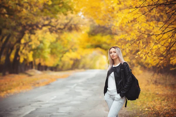 郊外の道路のアスファルトの道路に立っているバックパックでカジュアルスポーツウェアの若い女性,女の子は歩くと秋の自然を楽しむ旅 — ストック写真