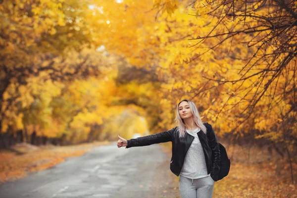 Junge Frau steht und wartet bei kaltem Herbstwetter auf einer asphaltierten Straße, ein Mädchen versucht, mit einer Handbewegung von einer Vorstadtautobahn zu kommen — Stockfoto